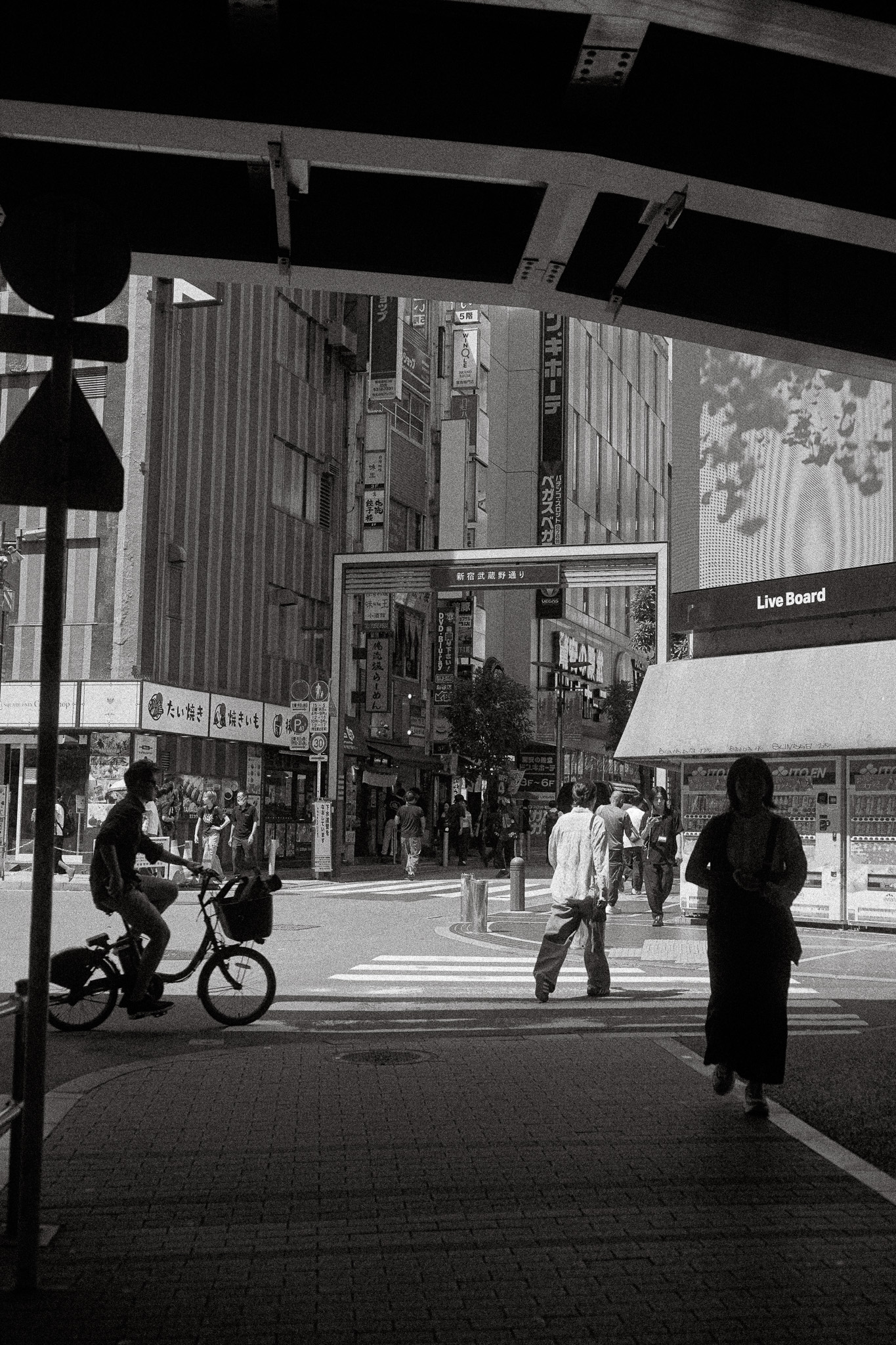 Black and white photo of a city street scene with a person riding a bicycle, pedestrians crossing a crosswalk, and tall buildings with Japanese shop signs.